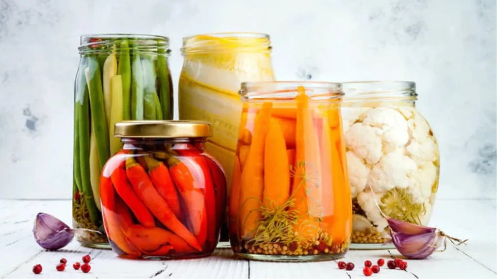 Glass jars filled with assorted fermented vegetables including carrots, cauliflower, peppers, and green beans on a white wooden surface.