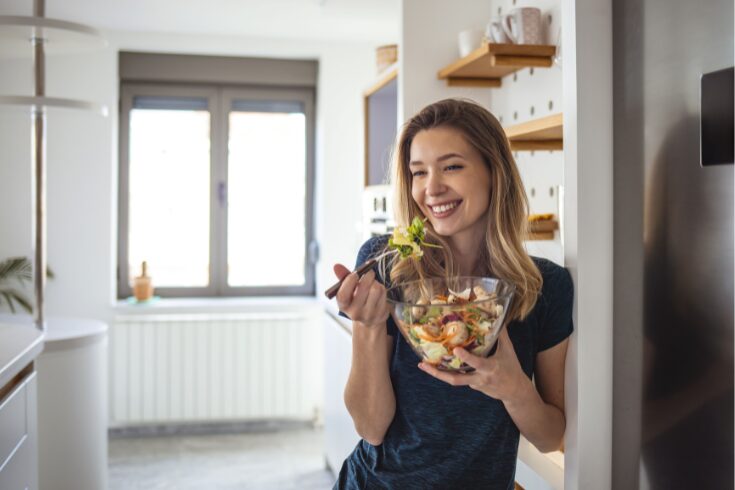 A woman in a kitchen holds a bowl of fresh salad, showcasing healthy ingredients and a vibrant meal preparation.
